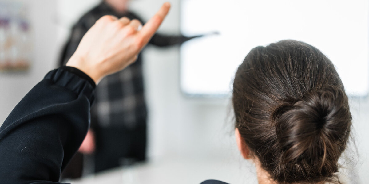 Eine Person meldet sich während ein Mann im Hintergrund etwas an einem Whiteboard zeigt.