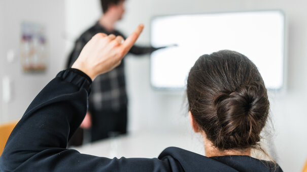Eine Person meldet sich während ein Mann im Hintergrund etwas an einem Whiteboard zeigt.