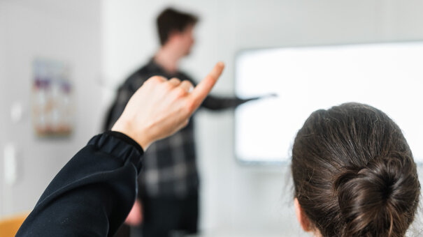 Eine Person meldet sich während ein Mann im Hintergrund etwas an einem Whiteboard zeigt.