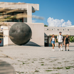 Eine Kunstwerk aus Bronze in Form einer großen Kugel steht vor dem Zentralen Hörsaalgebäude der Universität Regensburg. 