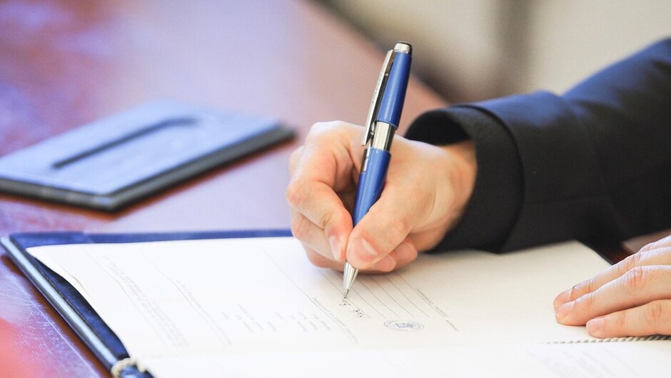 Zu sehen ist ein Schreibtisch. Auf diesem liegt ein Papierdokument mit Siegel, das von einer Person mit Kugelschreiber unterschrieben wird. 