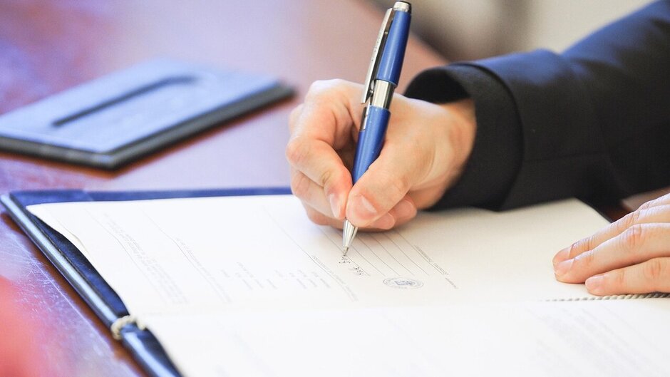 Zu sehen ist ein Schreibtisch. Auf diesem liegt ein Papierdokument mit Siegel, das von einer Person mit Kugelschreiber unterschrieben wird. 