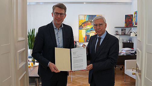 Zwei Männer in formeller Kleidung stehen in einer geöffneten Flügeltür, die den Blick auf ein Büro freigibt. Beide Männer halten gemeinsam eine geöffnete Dokumentenmappe mit einem förmlichen Schreiben darin in Richtung Kamera.