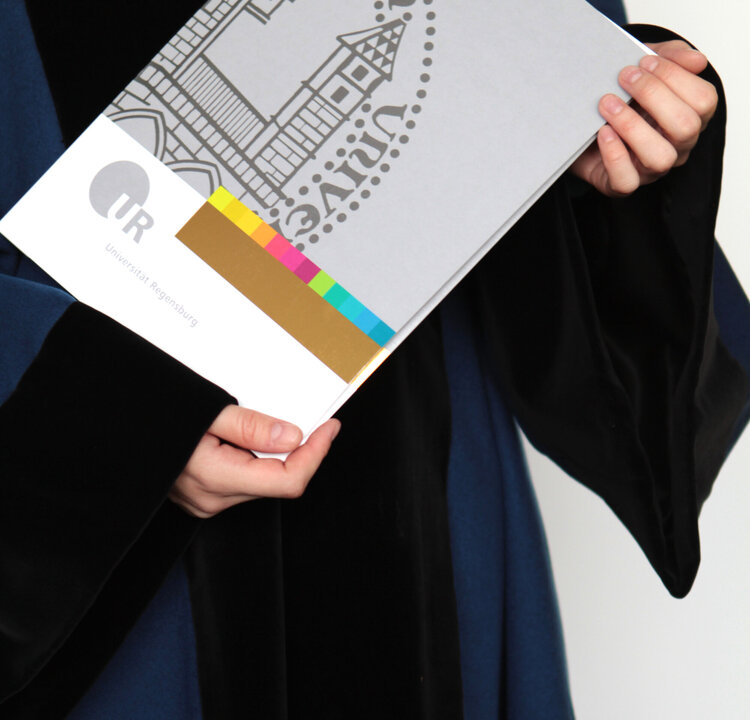 Eine Person im Talar hält eine Urkundenmappe mit dem Siegel der Universität Regensburg in den Händen.