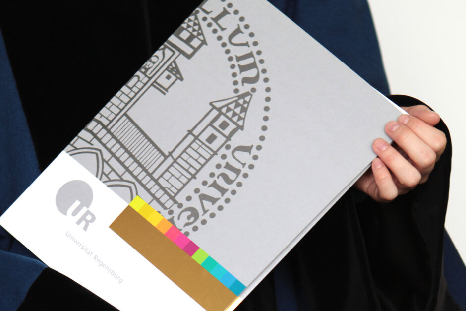 Eine Person im Talar hält eine Urkundenmappe mit dem Siegel der Universität Regensburg in den Händen.