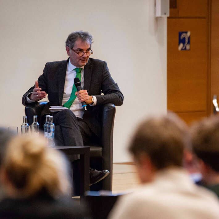 Ein älterer Mann mit grau melierten Haaren, Brille, Anzug und grüner Krawatte sitzt auf einem schwarzen Ledersessel und spricht in ein Mikrofon, das er in der Hand hält. (öffnet Vergrößerung des Bildes)