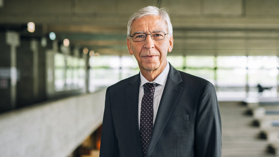 Ein Mann im Anzug und mit Krawatte, mit grauem Kurzhaarschnitt und Brille, steht entspannt und dem Betrachter zugewandt in einem weitläufigen Gebäude mit vielen Fensterfronten.