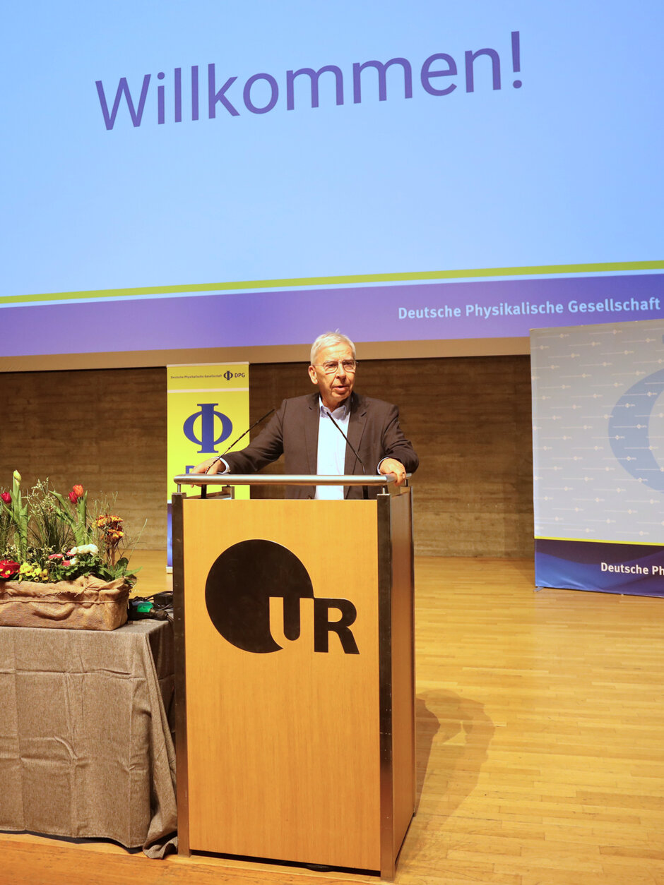 Ein älterer Mann im Anzug steht hinter einem mit UR-Logo geschmückten Rednerpult. Hinter ihm, auf einer Leinwand, ist ein großes "Willkommen" projiziert.