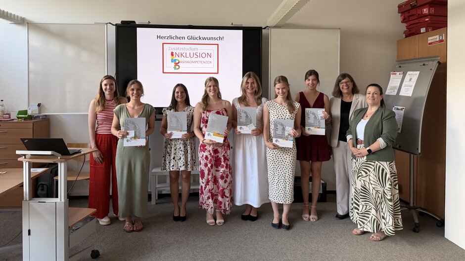 Neun Personen stehen in einem Seminarraum vor einem Bildschirm und halten Zertifikatsmappen in den Händen. Auf dem Bildschirm ist die Aufschrift „Herzlichen Glückwunsch!“ sowie das Logo des „Zusatzstudiums Inklusion Basiskompetenzen“ zu sehen. Der Raum ist mit Whiteboards, einem mobilen Präsentationsbildschirm und Unterrichtsmobiliar ausgestattet. Die Gruppe ist für ein gemeinsames Erinnerungsfoto nach einer Zertifikatsverleihung aufgestellt. (öffnet Vergrößerung des Bildes)