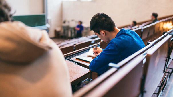 Student, der von hinten zu sehen ist, sitzt in einem hellen Hörsaal. Er schreibt etwas auf seinen Block.