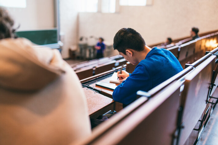 Student, der von hinten zu sehen ist, sitzt in einem hellen Hörsaal. Er schreibt etwas auf seinen Block. (opens enlarged image)