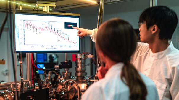 Zwei Personen in weißen Laborkitteln stehen vor einem physikalischen Versuchsaufbau und betrachten ein Diagramm auf einem Bildsschirm.