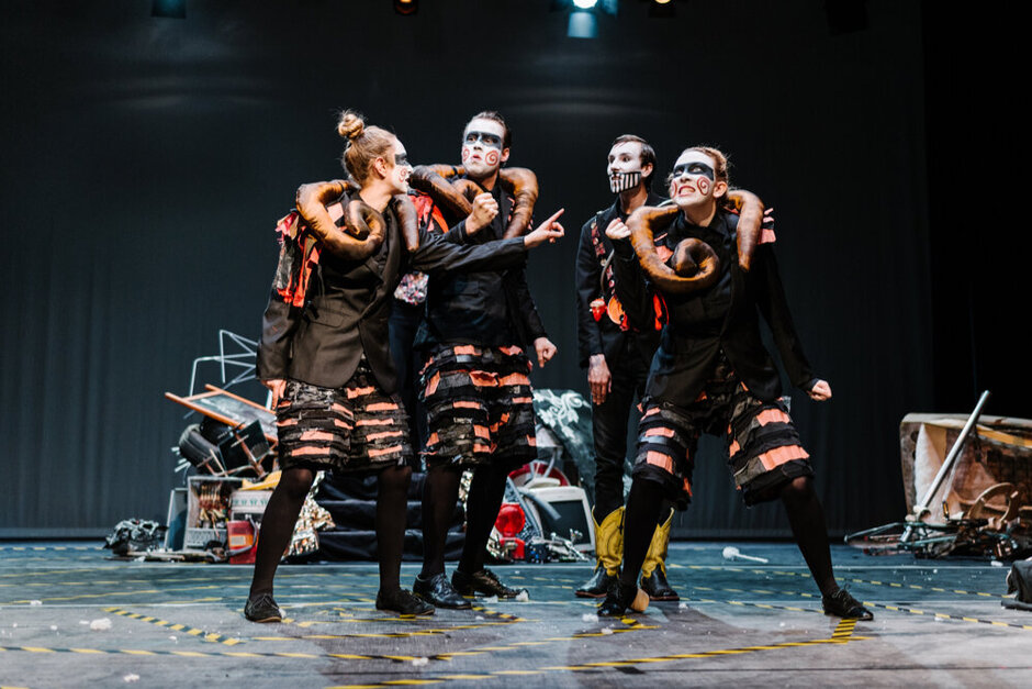 Vier junge Personen in dunklen Kostümen und mit bemalten Gesichtern stehen mit ausdrucksstarker Mimik auf einer Theaterbühne.