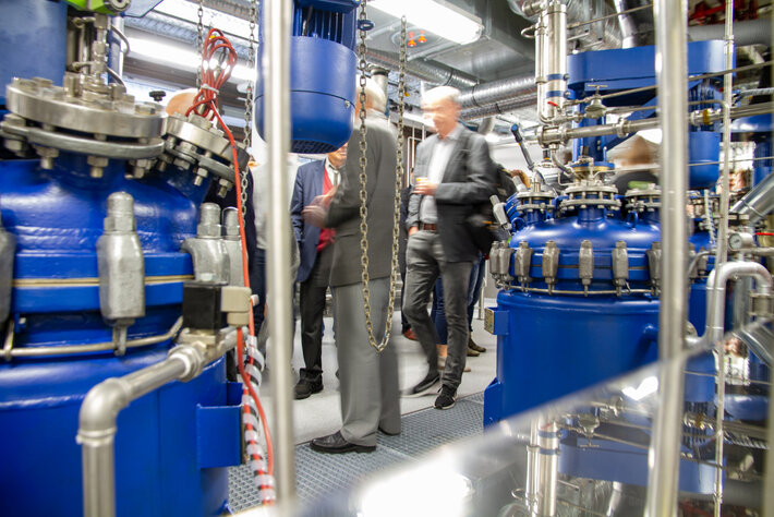 Ein hell erleuchteter Laborraum, in dem blau gestrichene Metalltanks und silbern glänzende Leitungen zu sehen sind. In der Bildmitte bewegen sich Personen in formeller Kleidung durch den Raum. (öffnet Vergrößerung des Bildes)