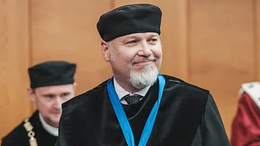Person in schwarzer akademischer Amtstracht mit Barett und Medaille an blauem Band hält eine blaue Urkundenmappe. Die Person steht in einem holzvertäfelten Saal, im Hintergrund sind weitere Personen in akademischer Kleidung zu sehen. (öffnet Vergrößerung des Bildes)