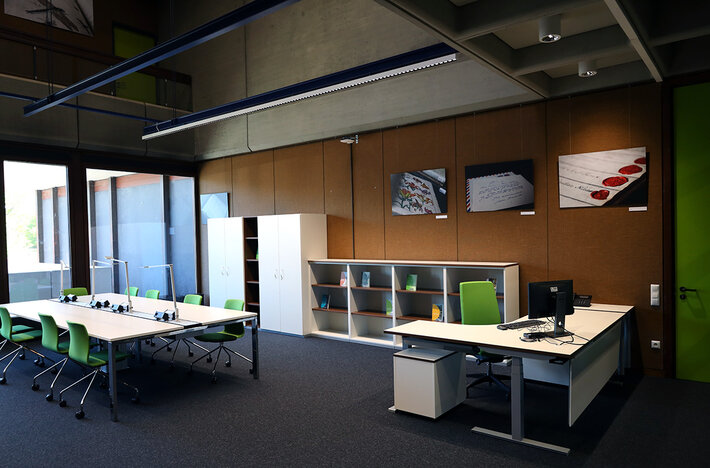 Arbeitsplätze, Schreibtisch mit Stuhl und Bildschirm, im Hintergrund Regal mit Büchern und Wandbilder (öffnet Vergrößerung des Bildes)