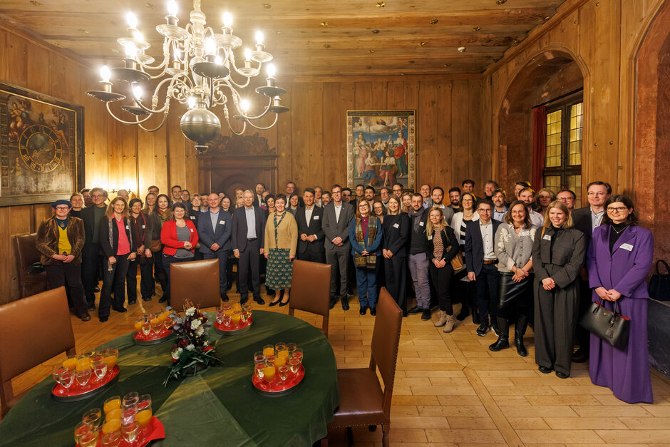 Rund 70 Personen stehe im Halbkreis im Kurfürstenzimmer des Alten Rathauses in Regensburg. Das Zimmer ist vollständig holzvertäfelt. Im Vordergrund steht ein großer runder Tisch mit den noch unbenutzten Gläsern eines Sektempfangs. (öffnet Vergrößerung des Bildes)