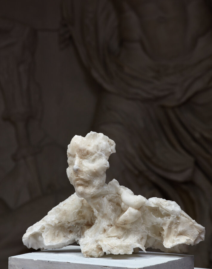 Eine abstrakte Wachsskulptur: Ein menschlicher Torso mit angedeuteten Gesichtszügen und rauer, unvollständiger Oberfläche. Die Skulptur steht vor einem unscharfen Hintergrund mit drapiertem Stoff. (öffnet Vergrößerung des Bildes)