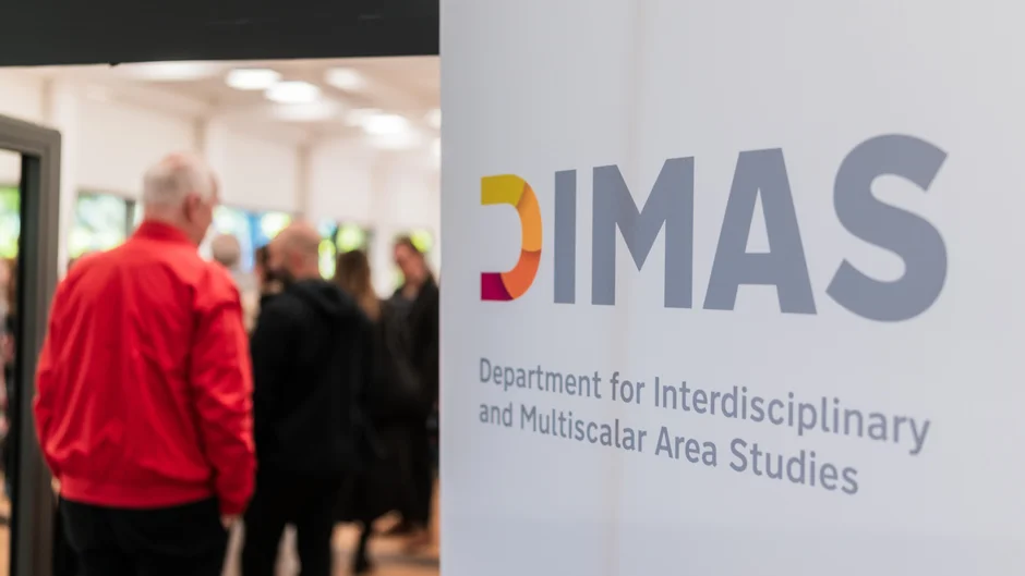 On the right-hand side is a roll-up banner with the DIMAS logo. To the left of it, several people can be seen blurred at the entrance to a seminar room, talking to each other.