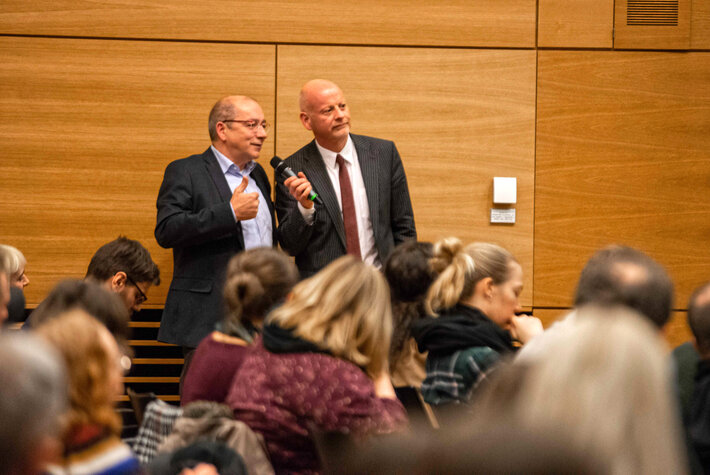Zwei Männer in förmlicher Kleidung stehen am Rande eines Hörsaals. Der eine Mann hält dem anderen ein Mikrofon hin. (öffnet Vergrößerung des Bildes)