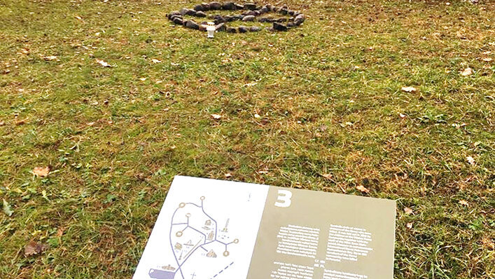 Eine von hohen Bäumen umstandene Wiese, in der Mitte ein Steinkreis. Im Vordergrund ist eine Informationstafel mit einem Lageplan und einem Textblock zu sehen.