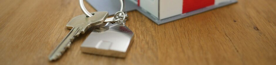 Kleines Häuschen in Spielzeuggröße auf einem Tisch, davor ein Schlüssel mit Anhänger