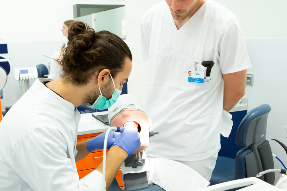 Eine Person mit einer medizinischen Gesichtsmaske, Laborkittel und blauen Einmalhandschuhen hantiert mit einem Gerät an einem Dummy-Kopf.