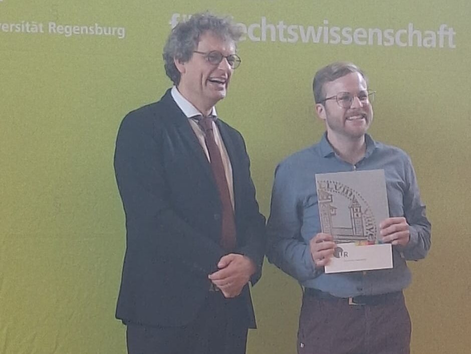 Prof. Graser überreicht Zachariasz Hussendörfer die Urkunde auf der Bühne vor einem Aufsteller mit dem Logo der Fakultät. (öffnet Vergrößerung des Bildes)