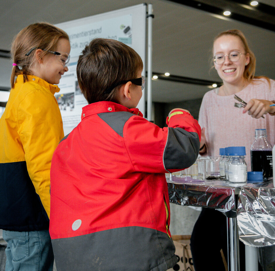 Zwei Kinder stehen fasziniert am Stand einer jungen Wissenschaftlerin, die chemische Experimente zum Mitmachen anbietet.