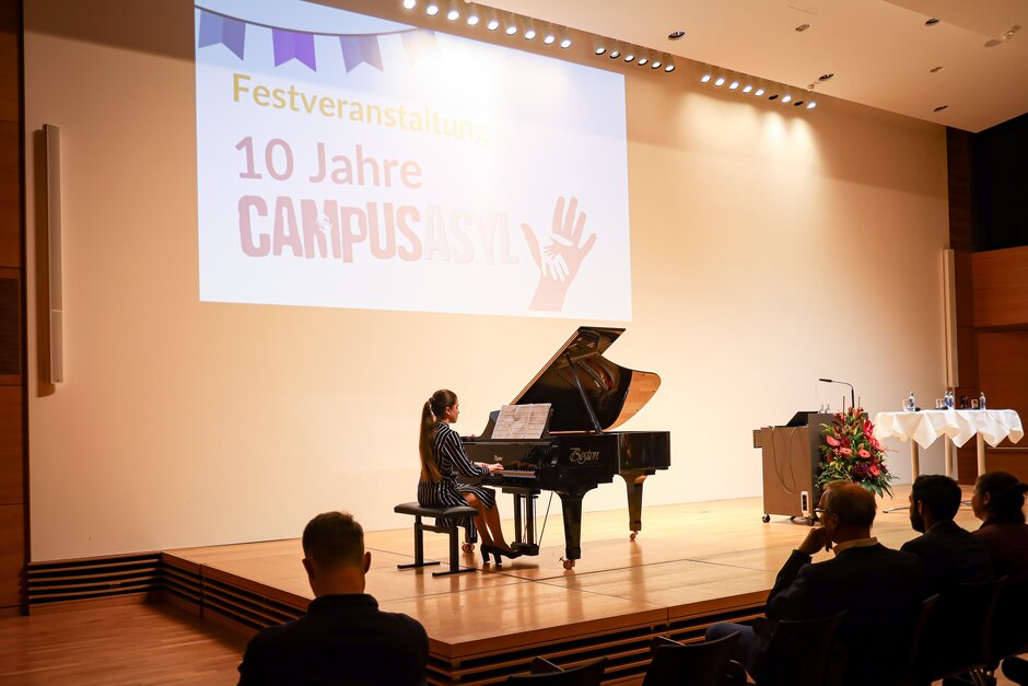 Frau sitzend an Flügel; im Hintergrund Projektion "Festveranstaltung 10 Jahre CampusAsyl"