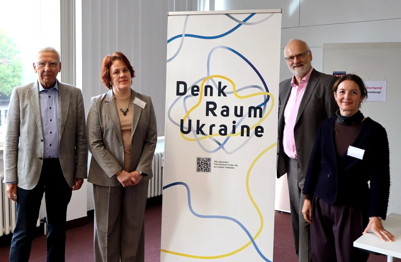 Drei Personen stehen neben einem Banner mit der Aufschrift „DenkRaum Ukraine“ in einem hellen Innenraum. Das Banner zeigt geschwungene Linien in Blau, Grau und Gelb sowie einen QR-Code. (öffnet Vergrößerung des Bildes)
