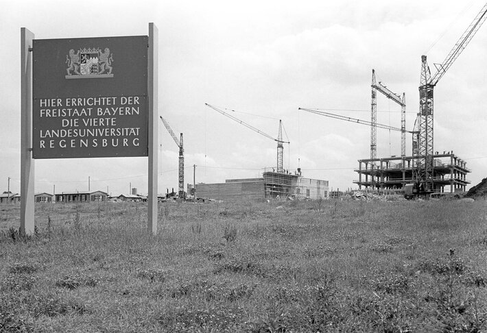 Das schwarz-weiß Foto zeigt eine Wiese mit einer Bautafel auf der steht: "Hier errichtet der Freistaat Bayern die Vierte Landesuniversität Regensburg". Im Hintergrund sind Baukräne und Hörsaalgebäude im Rohbau zu erkennen. (opens enlarged image)