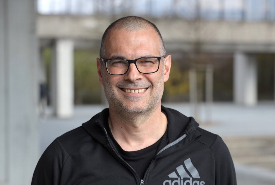 Porträtbild: Mann mit schwarzer Sportjacke, schwarzer Brille und sehr kurzen, dunklen Haaren, im freien aufgenommen - im Hintergrund angerissen Teile des UR-Rechenzentrum-Gebäudes