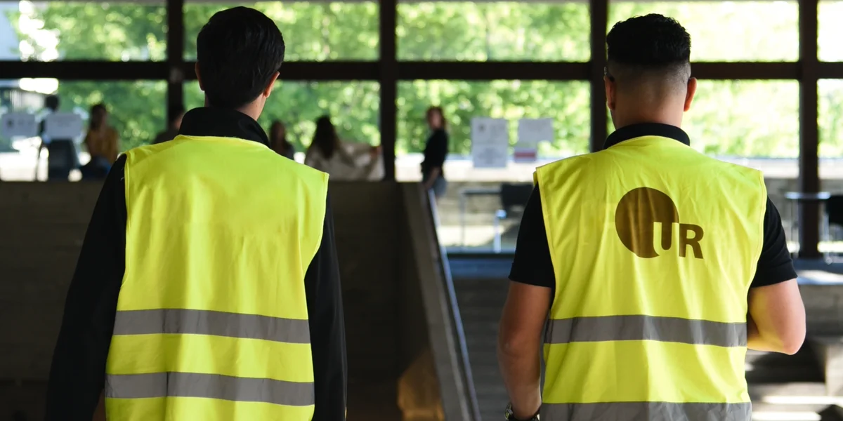 Zwei Männer mit gelben Warnwesten im Zentralen Hörsaalgebäude. Auf einer Warnweste ist das UR-Logo der Universität Regensburg angebracht.