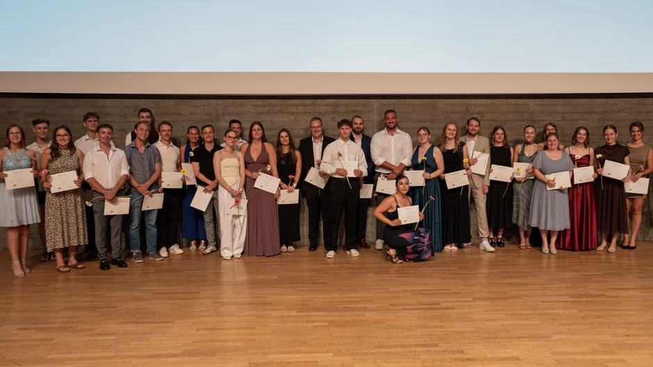 Gruppenfoto auf einer Bühne mit Holzfußboden vor einer Betonwand. Rund 30 Personen stehen in mehreren Reihen und halten Urkunden sowie einzelne Blumen in den Händen. Die Teilnehmenden tragen festliche Kleidung. Im Hintergrund ist eine große Projektionsfläche zu sehen. (öffnet Vergrößerung des Bildes)