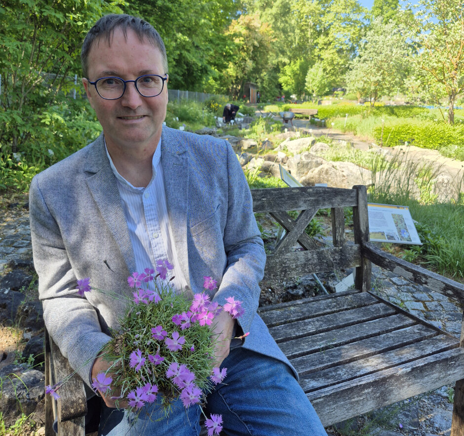 Ein Mann sitzt auf einer Holzbank und hält rosa blühende Pflanzen in der Hand. Im Hintergrund ist eine parkähnliche Gartenanlage zu sehen.