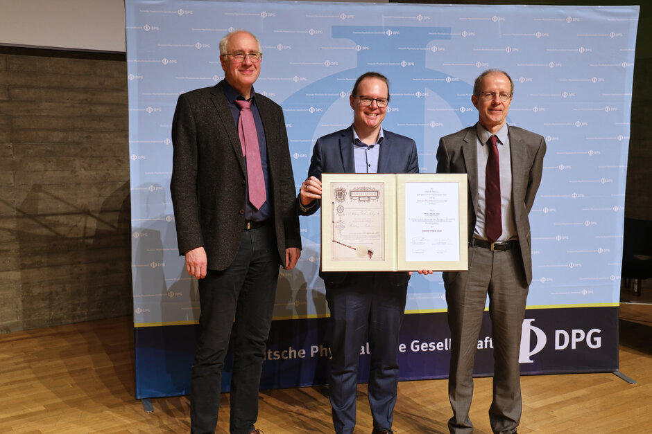 Drei Männer in Anzügen stehen vor einer mit DPG-Logo bedruckten Fläche. Der mitllere Mann hält lächelnd eine Urkunde Richtung Kamera.