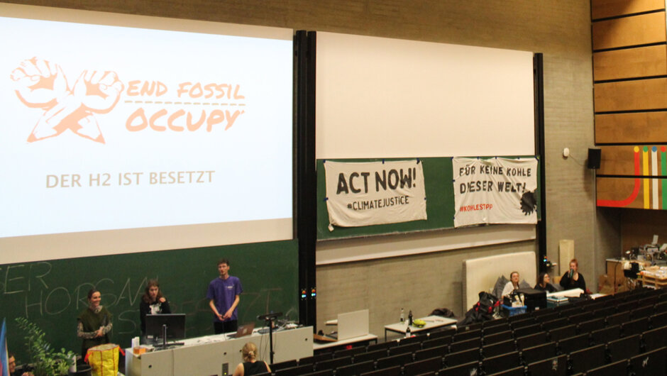 Ein Hörsaal mit großer Leinwand, auf der die Texte ‚END FOSSIL OCCUPY‘ und ‚DER H2 IST BESETZT‘ angezeigt werden. An der Wand hängen zwei Banner: eines mit der Aufschrift ‚ACT NOW! #CLIMATEJUSTICE‘ und ein weiteres mit ‚FÜR KEINE KOHLE DIESER WELT #KOHLSTOP‘. Mehrere Personen befinden sich im Raum, einige stehen vorne in der Nähe eines Tisches.