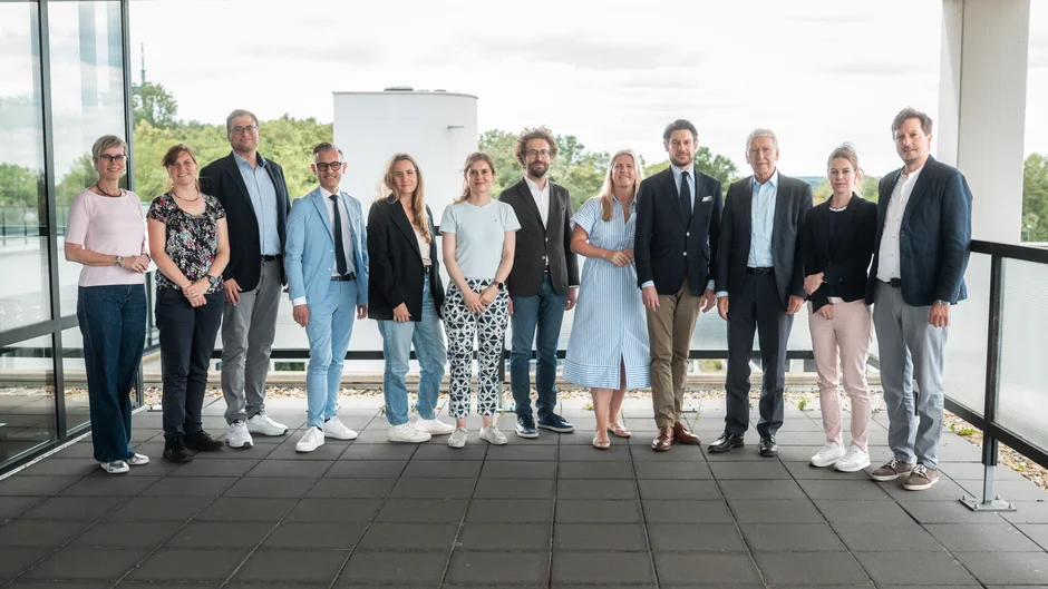 Gruppenfoto von zwölf Personen auf einer überdachten Terrasse vor einem modernen Gebäude. Im Hintergrund sind Bäume und ein bewölkter Himmel zu sehen. (öffnet Vergrößerung des Bildes)