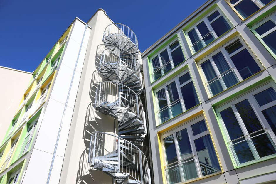 Eine Häuserfassade im Sonnenschein; zu sehen sind große Glasfenster mit integrierten französischen Balkonen und bunten Sonnnenblenden und eine Wendeltreppe aus Metall.