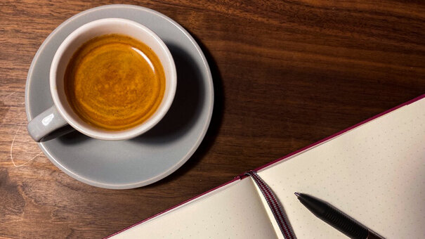 Eine Tasse Kaffee steht auf einer Oberfläche aus Holz, daneben liegt ein aufgeschlagenes Notizbuch mit Lesebändchen und Kugelschreiber.