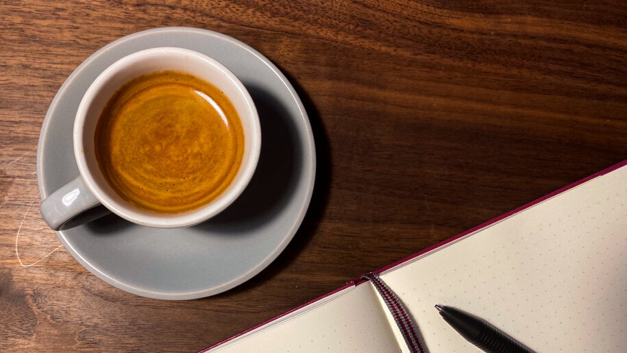 Eine Tasse Kaffee steht auf einer Oberfläche aus Holz, daneben liegt ein aufgeschlagenes Notizbuch mit Lesebändchen und Kugelschreiber.