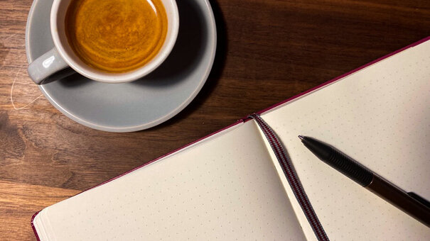 Eine Tasse Kaffee steht auf einer Oberfläche aus Holz, daneben liegt ein aufgeschlagenes Notizbuch mit Lesebändchen und Kugelschreiber.