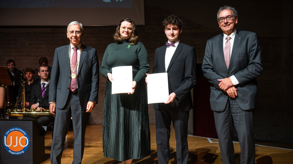 Vier Personen stehen auf einer Bühne und posieren für ein Foto. Zwei Personen in der Mitte halten Auszeichnungsurkunden in den Händen. Alle tragen formelle Kleidung. Im Hintergrund sitzt ein Ensemble mit Musikinstrumenten, und links ist ein Logo des Uni-Jazzorchesters sichtbar.