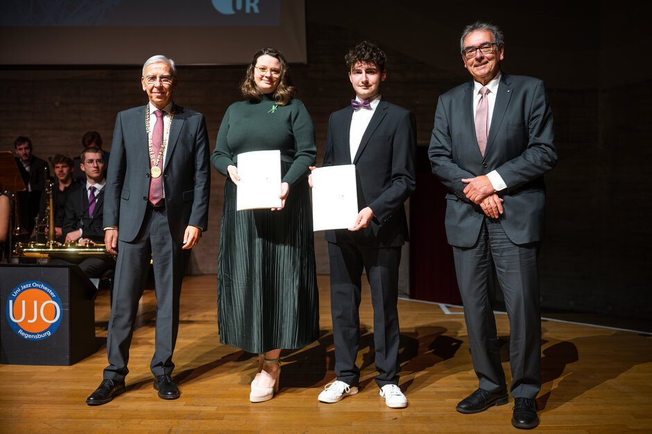Vier Personen stehen auf einer Bühne und posieren für ein Foto. Zwei Personen in der Mitte halten Auszeichnungsurkunden in den Händen. Alle tragen formelle Kleidung. Im Hintergrund sitzt ein Ensemble mit Musikinstrumenten, und links ist ein Logo des Uni-Jazzorchesters sichtbar.