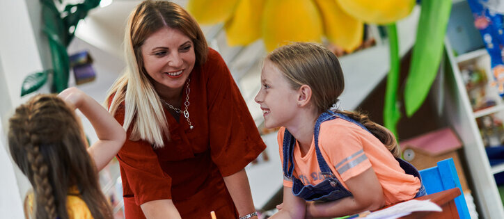 Zwei Kinder sitzen einander gegenüber an einem Tisch, vor ihnen liegen Stifte und Papier. Zwischen Ihnen steht eine Lehrerin und erklärt etwas. Alle Personen haben Stifte in der Hand.