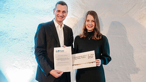 Ein Herr in dunklem Anzug und weißem Hemd hält eine Urkunde in die Kamera. Neben ihm eine schwarz gekleidete junge Frau mit schulterlangem Haar, die einen Dekoscheck hält.