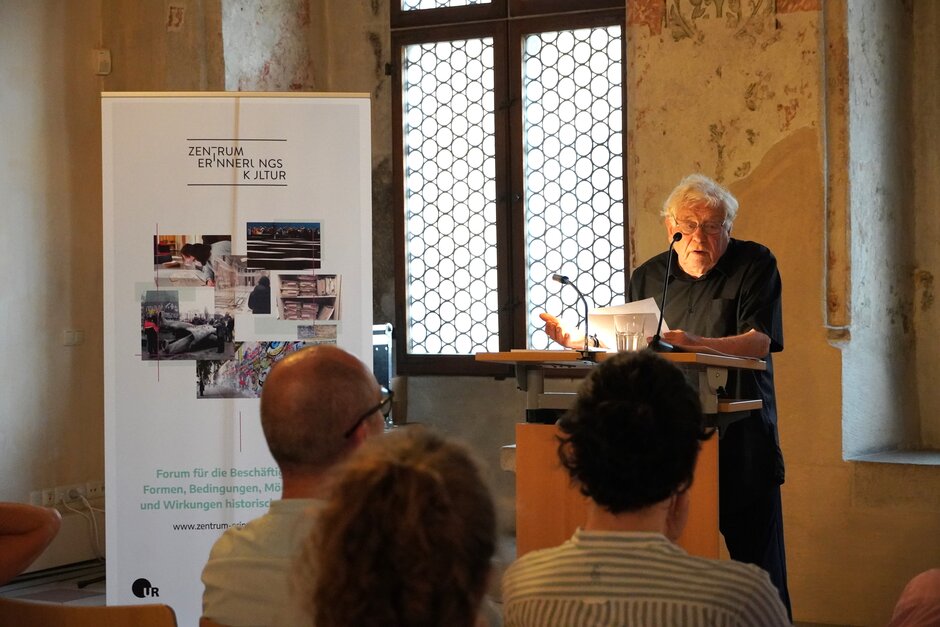 Älterer Herr an Rednerpult, spricht zu Publikum.