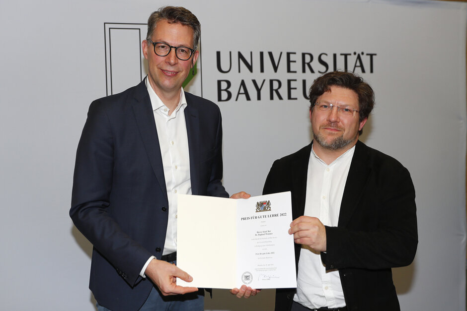 Zwei Männer in formeller Kleidung schauen in Richtung des Betrachters und halten gemeinsam eine geöffente Urkundenmappe. Auf dem Dokument in der Mappe ist das Bayerische Staatswappen zu erkennen. (öffnet Vergrößerung des Bildes)