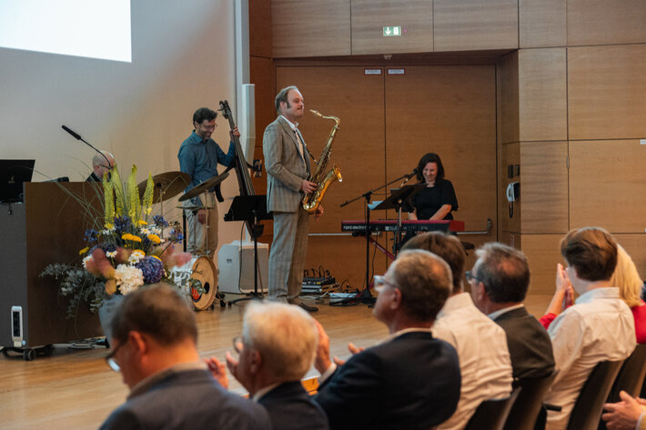 Ein Schlagzeuger, ein Bassist, ein Saxophonspieler und eine Pianistin sind mit ihren Instrumenten auf einer mit Blumen geschmückten Bühne. (öffnet Vergrößerung des Bildes)